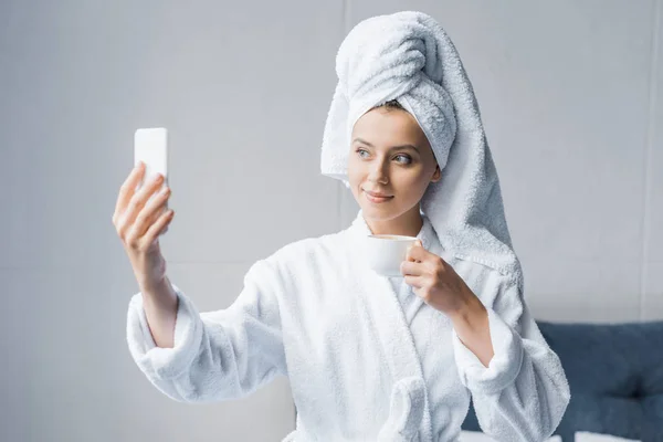 Belle fille en peignoir et serviette prendre selfie avec une tasse de café le matin — Photo de stock