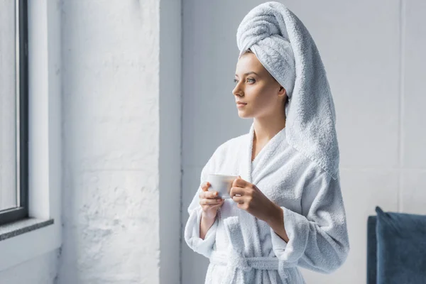 Mujer joven en albornoz y toalla blanca en la cabeza sosteniendo taza de café y mirando a la ventana - foto de stock