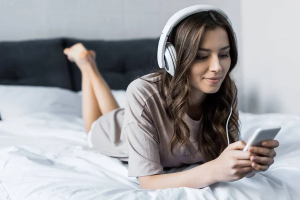 Bela morena menina ouvir música com fones de ouvido e smartphone enquanto deitado na cama — Fotografia de Stock