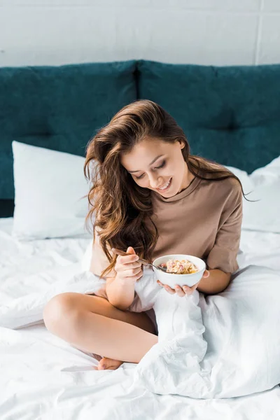 Glückliches Mädchen isst Haferflocken zum Frühstück, während es auf dem Bett sitzt — Stockfoto