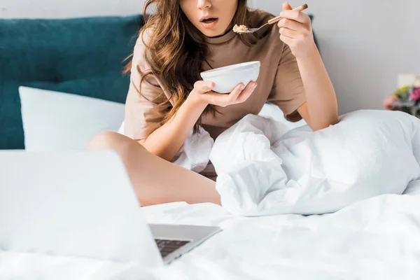 Обрізаний вид шокованої дівчини, що їсть її сніданок і дивиться щось на ноутбук, сидячи на ліжку — стокове фото