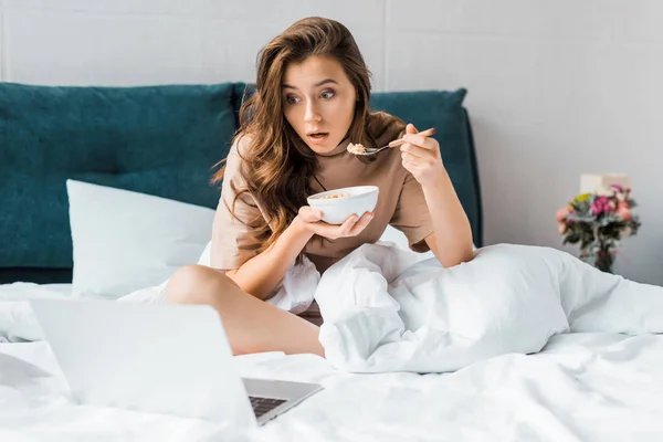 Shocked brunette girl with bowl of oatmeal watching something on laptop while sitting on bed — Stock Photo