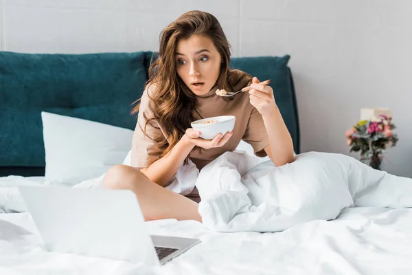 Choqué fille brune avec bol de flocons d'avoine regarder quelque chose sur ordinateur portable tout en étant assis sur le lit — Photo de stock