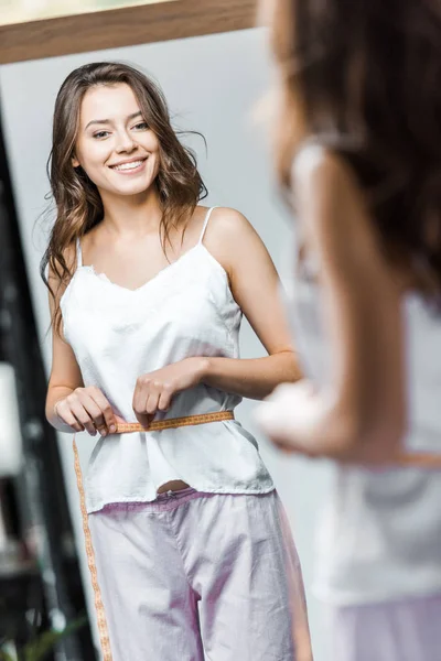 Souriant jeune femme mesurant sa taille et regardant miroir — Photo de stock