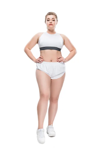 Full length view of young size plus woman in sportswear standing with hands on waist and looking at camera isolated on white — Stock Photo