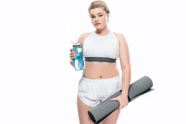 Atractiva chica con sobrepeso en ropa deportiva celebración botella de agua y esterilla de yoga aislado en blanco - foto de stock