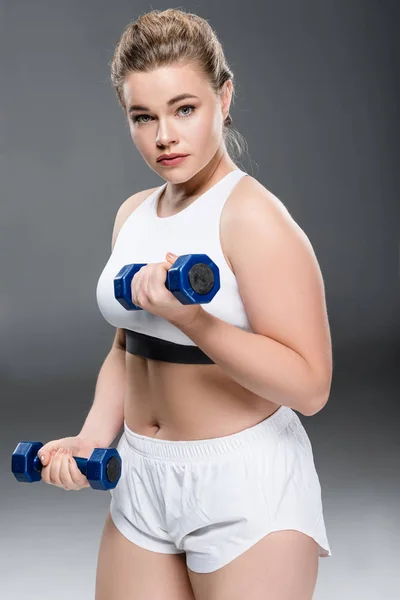 Jeune taille plus femme faisant de l'exercice avec haltères et regardant la caméra sur gris — Photo de stock