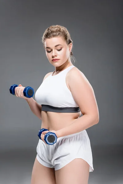 Jeune taille plus femme faisant de l'exercice avec haltères et regardant vers le bas sur gris — Photo de stock