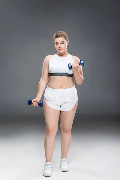 Jeune femme oversize en vêtements de sport faisant de l'exercice avec des haltères et regardant la caméra sur gris — Photo de stock