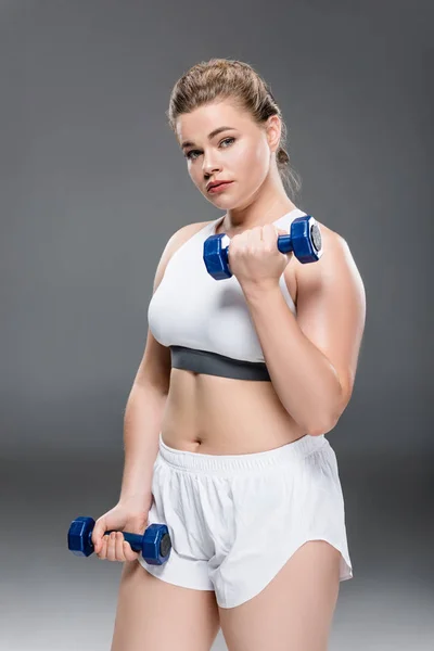 Jeune femme en surpoids en vêtements de sport faisant de l'exercice avec des haltères et regardant la caméra sur gris — Photo de stock