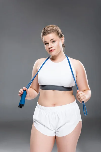 Joven mujer de gran tamaño en ropa deportiva sosteniendo saltar la cuerda y mirando a la cámara en gris - foto de stock