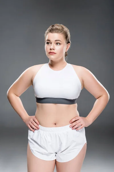 Atractiva joven mujer de gran tamaño en ropa deportiva de pie con las manos en la cintura y mirando hacia otro lado en gris - foto de stock