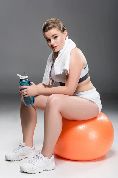 Joven mujer con sobrepeso con toalla y botella de agua sentada en la bola en forma y mirando a la cámara en gris - foto de stock