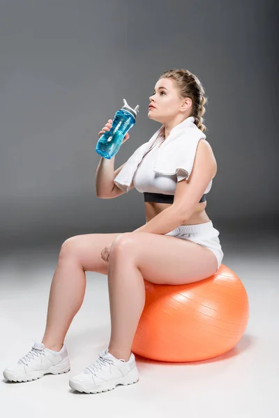 Young oversize woman with towel drinking water and sitting on fit ball on grey — Stock Photo