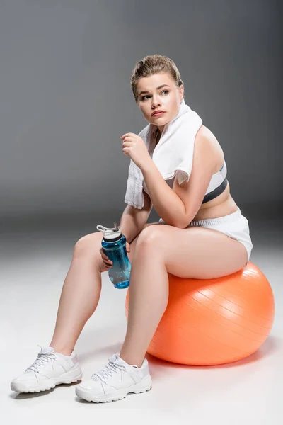 Young oversize woman with towel and bottle of water sitting on fit ball on grey — Stock Photo