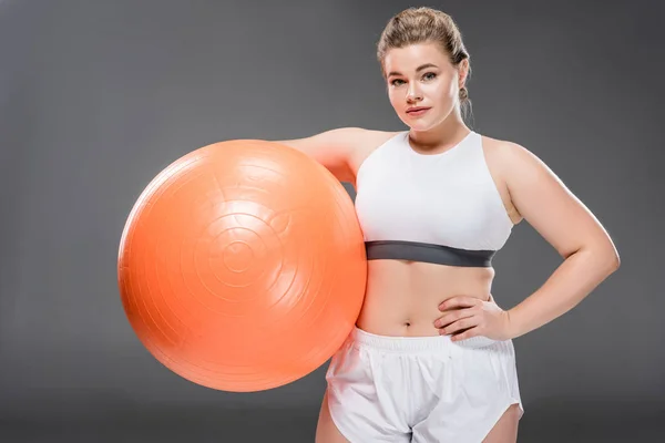 Junge übergewichtige Frau in Sportkleidung, die einen fitten Ball hält und in die Kamera schaut, isoliert auf grau — Stockfoto