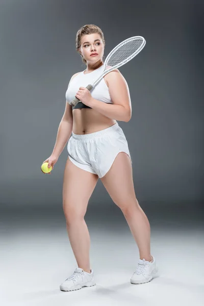 Joven mujer de gran tamaño jugando al tenis y mirando hacia otro lado en gris - foto de stock