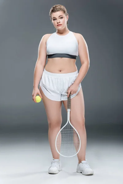 Young plus size woman in sportswear holding tennis racquet and ball on grey — Stock Photo