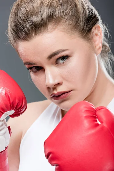 Vista de cerca de la joven mujer de gran tamaño en guantes de boxeo mirando a la cámara aislada en gris - foto de stock
