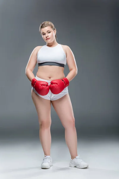 Visão de comprimento total da jovem mulher com sobrepeso em roupas esportivas e luvas de boxe olhando para a câmera em cinza — Fotografia de Stock