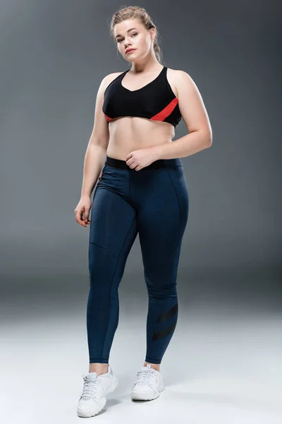 Ganzkörperansicht der jungen Plus-Size-Frau in Sportbekleidung mit Blick auf die Kamera auf grau — Stockfoto