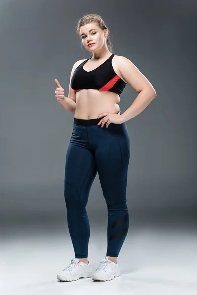 Hermosa chica con sobrepeso en ropa deportiva mostrando el pulgar hacia arriba y mirando a la cámara en gris - foto de stock