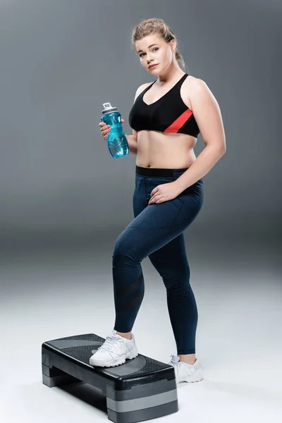 Joven mujer de gran tamaño en ropa deportiva celebración botella de agua y de pie en la plataforma de paso en gris - foto de stock