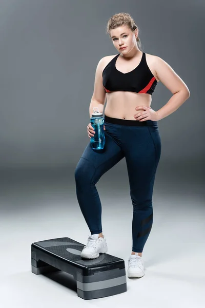 Young overweight woman in sportswear holding bottle of water and standing on step platform on grey — Stock Photo