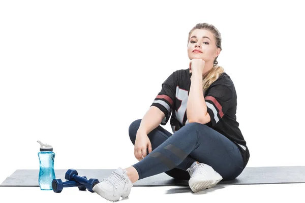 Young overweight woman in sportswear sitting on yoga mat and looking at camera isolated on white — Stock Photo
