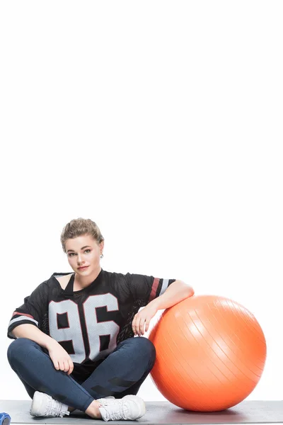 Jeune femme en surpoids en vêtements de sport penché au ballon en forme et assis sur un tapis de yoga isolé sur blanc — Photo de stock