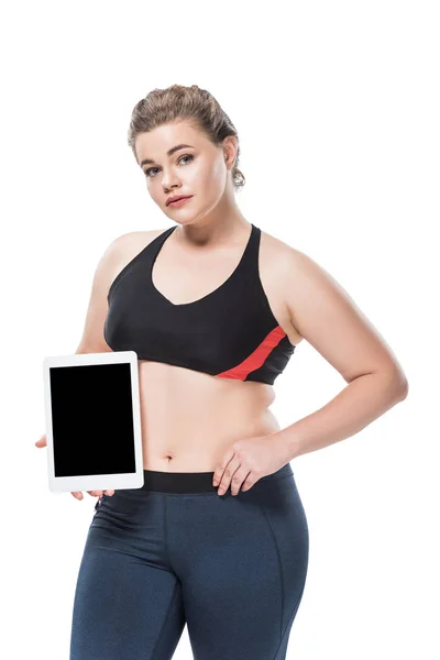 Young overweight woman in sportswear holding digital tablet with blank screen and looking at camera isolated on white — Stock Photo