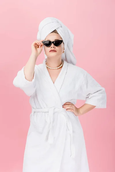 Jeune femme oversize en peignoir, lunettes de soleil et serviette sur la tête debout avec la main sur la taille isolé sur rose — Photo de stock