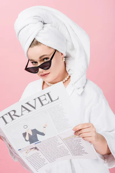 Mujer joven de gran tamaño en albornoz, gafas de sol y toalla en la cabeza leyendo el periódico de viaje y mirando a la cámara aislada en rosa - foto de stock