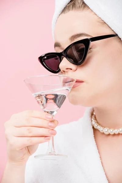 Vue rapprochée de jeune femme oversize en peignoir, lunettes de soleil et serviette sur la tête buvant du verre isolé sur rose — Photo de stock