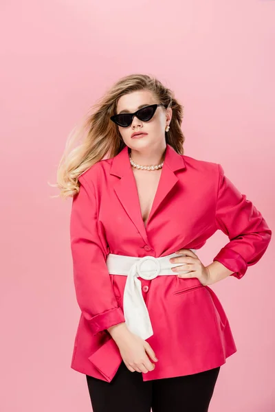 Attractive oversize girl in pink trench coat and sunglasses posing with hand on waist isolated on pink — Stock Photo