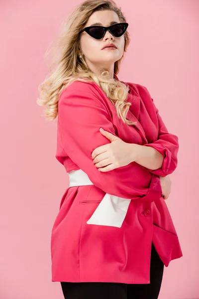 Attractive oversize girl in pink trench coat and sunglasses standing with crossed arms isolated on pink — Stock Photo