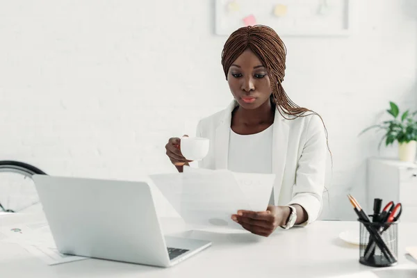 Atraente mulher de negócios afro-americana em terno branco sentado na mesa de computador, segurando xícara de café e trabalhando no projeto no escritório — Fotografia de Stock