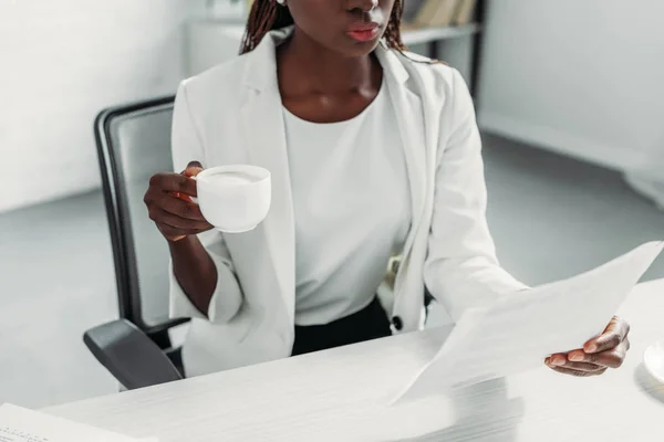 Sección media de la hermosa mujer de negocios adulta afroamericana en traje blanco sentada en el escritorio de la computadora, sosteniendo la taza de café y trabajando en la oficina - foto de stock