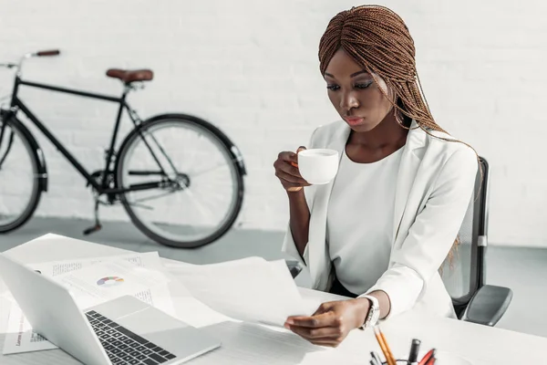 Mujer de negocios adulta afroamericana en traje blanco sentada en el escritorio de la computadora, sosteniendo la taza de café y trabajando en la oficina - foto de stock