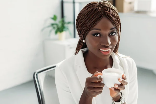Belle femme d'affaires africaine américaine adulte en costume blanc assis au bureau, tenant une tasse de café, souriant et regardant la caméra — Photo de stock