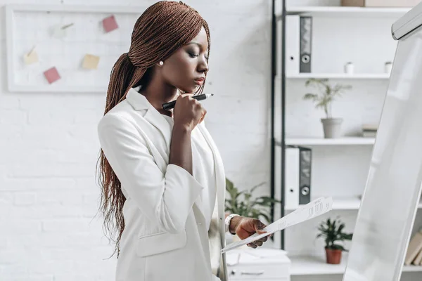 Nachdenkliche afrikanisch-amerikanische erwachsene Geschäftsfrau in weißer offizieller Kleidung hält Dokument in der Hand und arbeitet an einem Projekt in der Nähe von Flipchart im Büro — Stockfoto