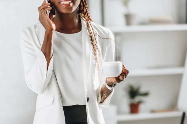 Середня частина афроамериканської дорослої бізнес-леді в білому костюмі розмовляє на смартфоні і тримає чашку кави в офісі — стокове фото