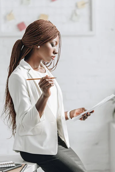 Focalizzata afroamericana donna d'affari adulta in bianco abbigliamento formale in possesso di matita, documento e lavoro in ufficio — Foto stock