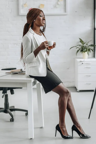 Africano americano adulto empresária em branco formal desgaste inclinado na mesa de escritório, sorrindo e segurando xícara de café — Fotografia de Stock