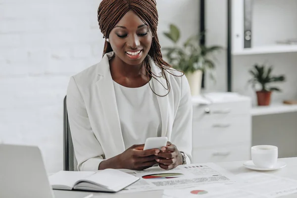 Sorridente donna d'affari afroamericana adulta seduta alla scrivania, che lavora e guarda lo smartphone — Foto stock