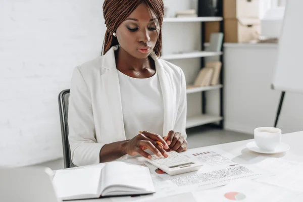 Focalizzata afroamericana donna d'affari adulta in abito formale bianco che lavora alla scrivania dell'ufficio e utilizza la calcolatrice — Foto stock