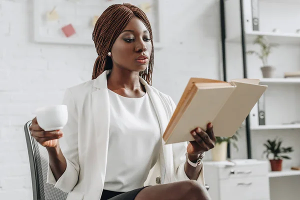 Grave afroamericana adulto mujer de negocios en blanco formal desgaste lectura libro y celebración de la taza de café en la oficina moderna - foto de stock