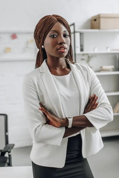 Selbstbewusste afrikanisch-amerikanische Geschäftsfrau in weißer offizieller Kleidung mit verschränkten Armen stehend und im Büro in die Kamera blickend — Stockfoto