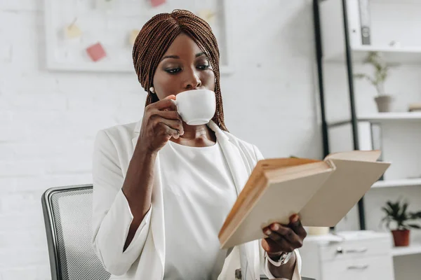 Africano americano adulto empresária em branco formal desgaste leitura livro e beber café no escritório — Fotografia de Stock