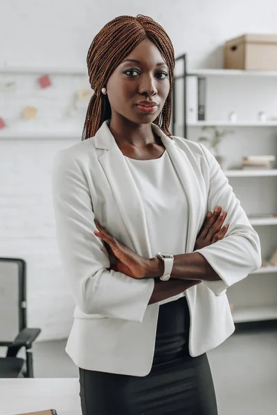 Ernst selbstbewusste afrikanisch-amerikanische Geschäftsfrau in weißer formeller Kleidung mit verschränkten Armen stehend und in die Kamera im Büro blickend — Stockfoto